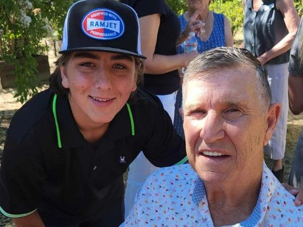 Young boy in with black hat and shirt and man in blue printed button up smiling together for a photo