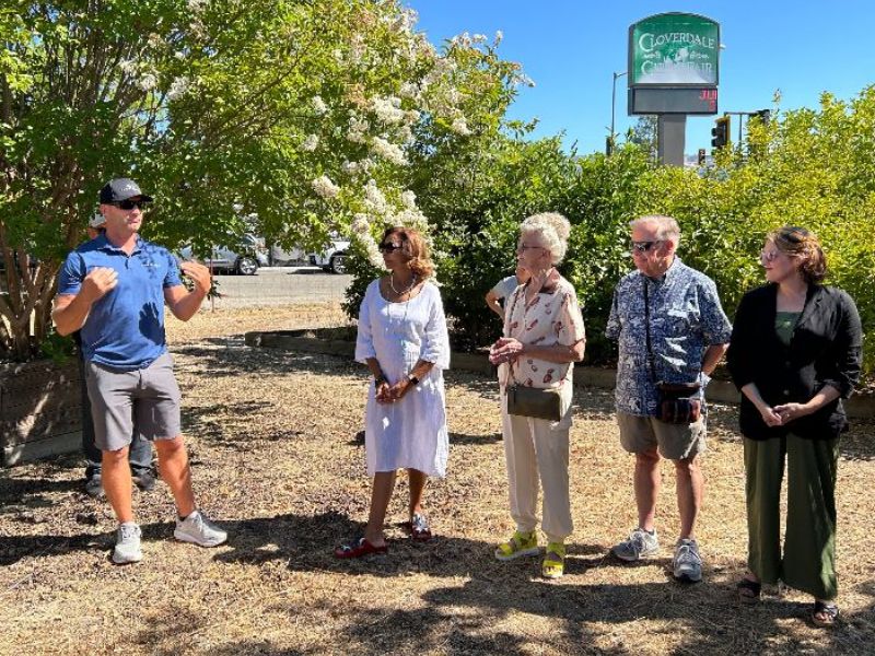 four people standing in front of trees looking and listening to a man speak